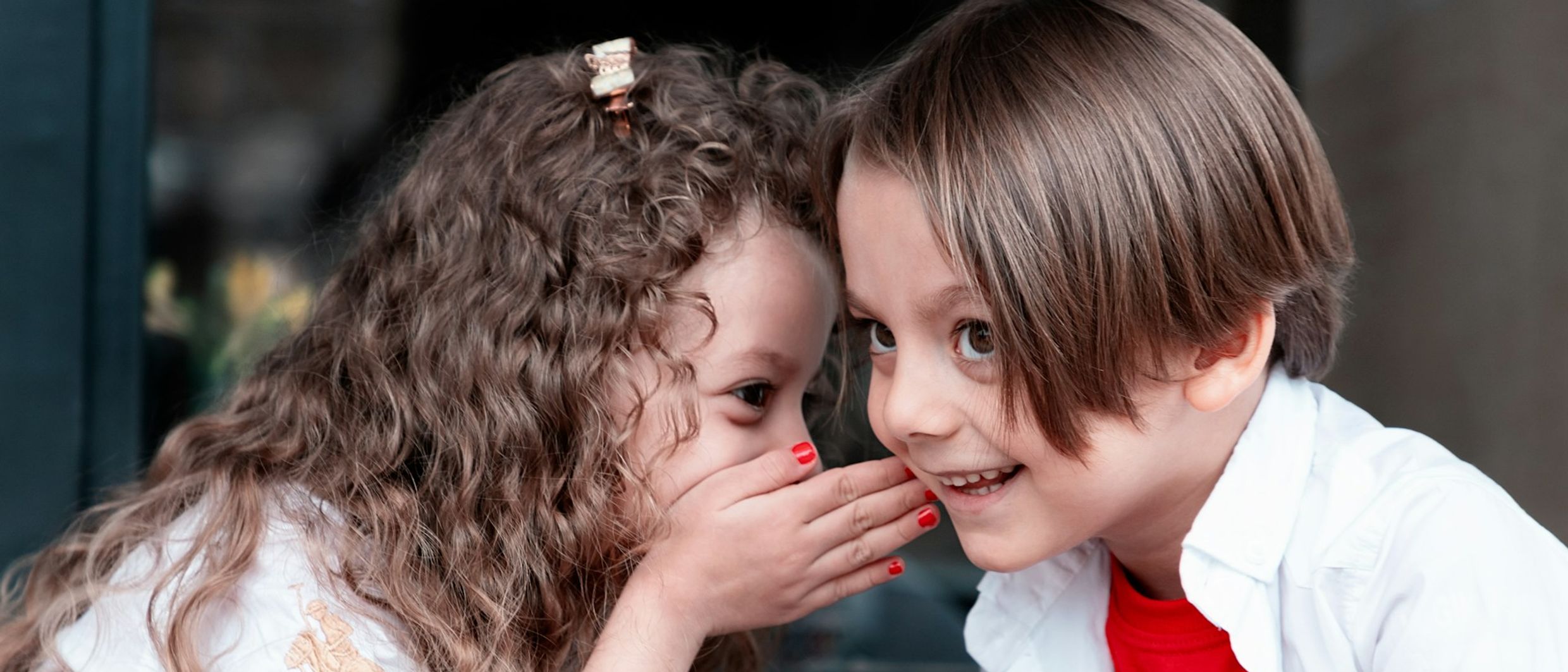 A young girl whispering a secret into a smiling boy’s ear