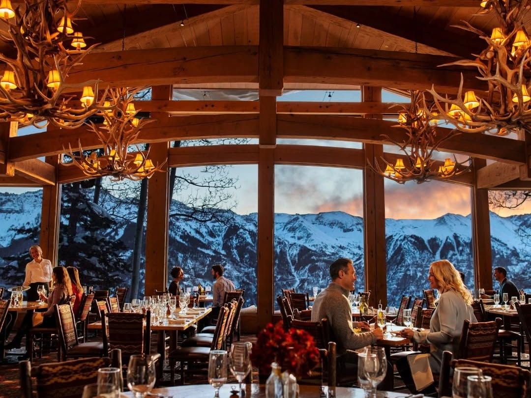 Elegant mountain-view dining room at sunset with guests enjoying meals under warm antler chandeliers. Floor-to-ceiling windows showcase snowcapped peaks, making it a signature Telluride dining experience.