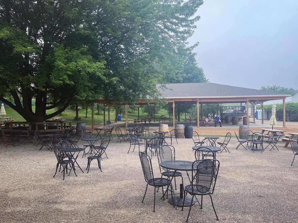 An empty outdoor patio at Lemon Creek Winery is filled with black metal bistro tables and chairs, wine barrels, and picnic tables beneath a large open-air pavilion. A misty morning fog softens the surrounding trees and greenery.