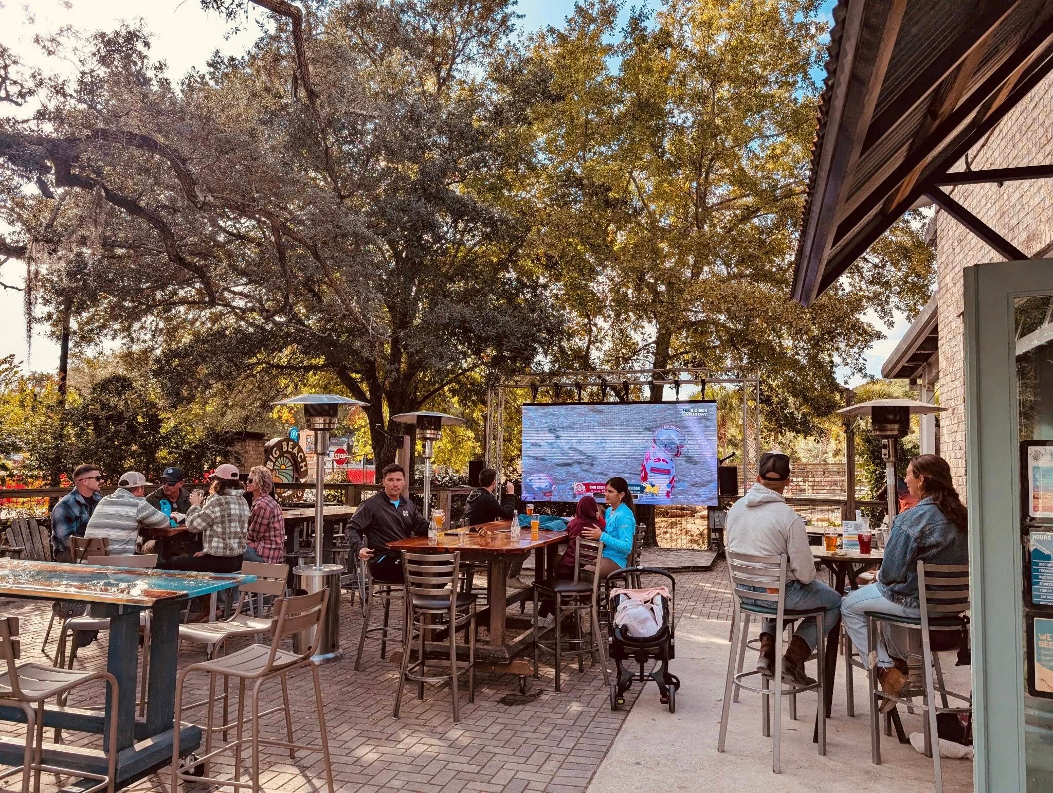 Outdoor patio at Big Beach Brewing Company with locals and visitors enjoying craft beer under shady trees. The relaxed setting, large screen, and open seating make it a popular spot to hang out, watch sports, and unwind with friends. It shows the casual brewery scene many travelers love when exploring the area.  File Name: