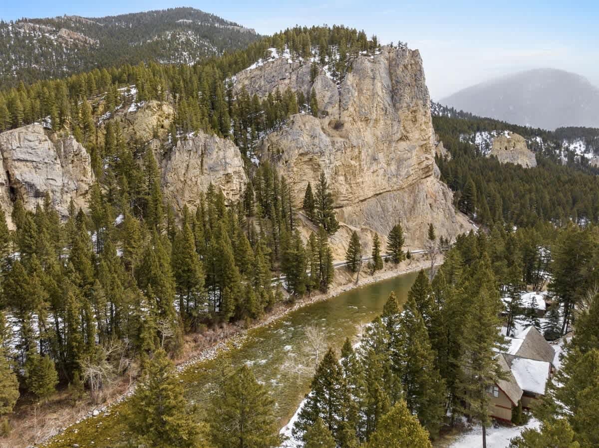 Towering cliffs rise above a winding river surrounded by pine forests in Big Sky, Montana. Light snow dusts the trees, creating a peaceful alpine setting perfect for outdoor adventures.