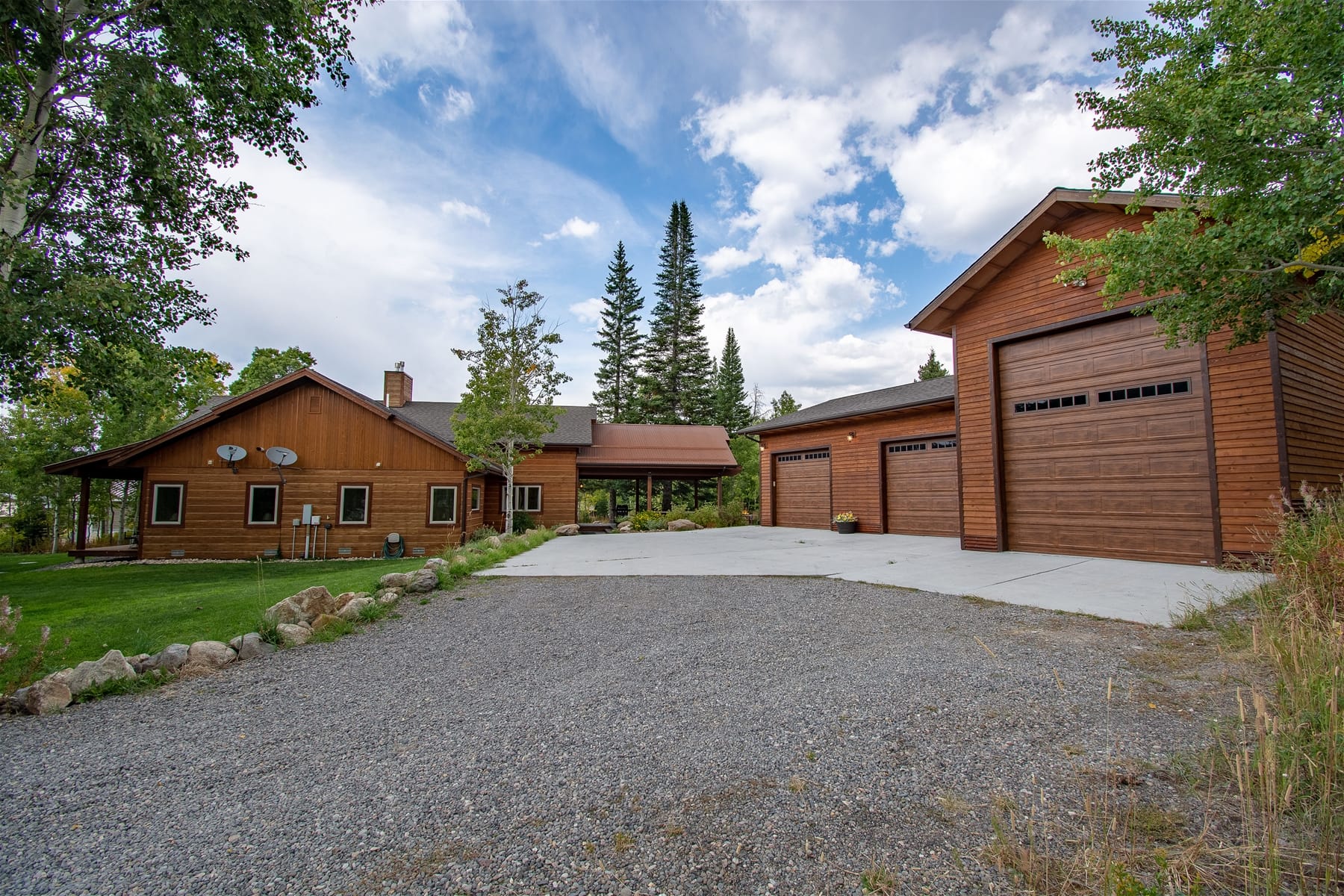 Rustic wooden home with a large detached three-door garage, set among trees with a gravel and concrete driveway under a partly cloudy sky.