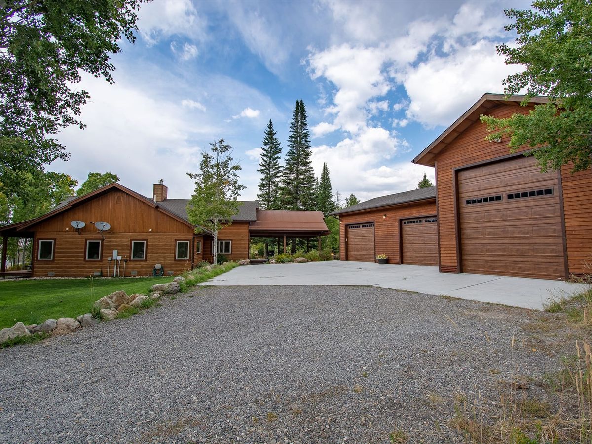 Rustic wooden home with a large detached three-door garage, set among trees with a gravel and concrete driveway under a partly cloudy sky.