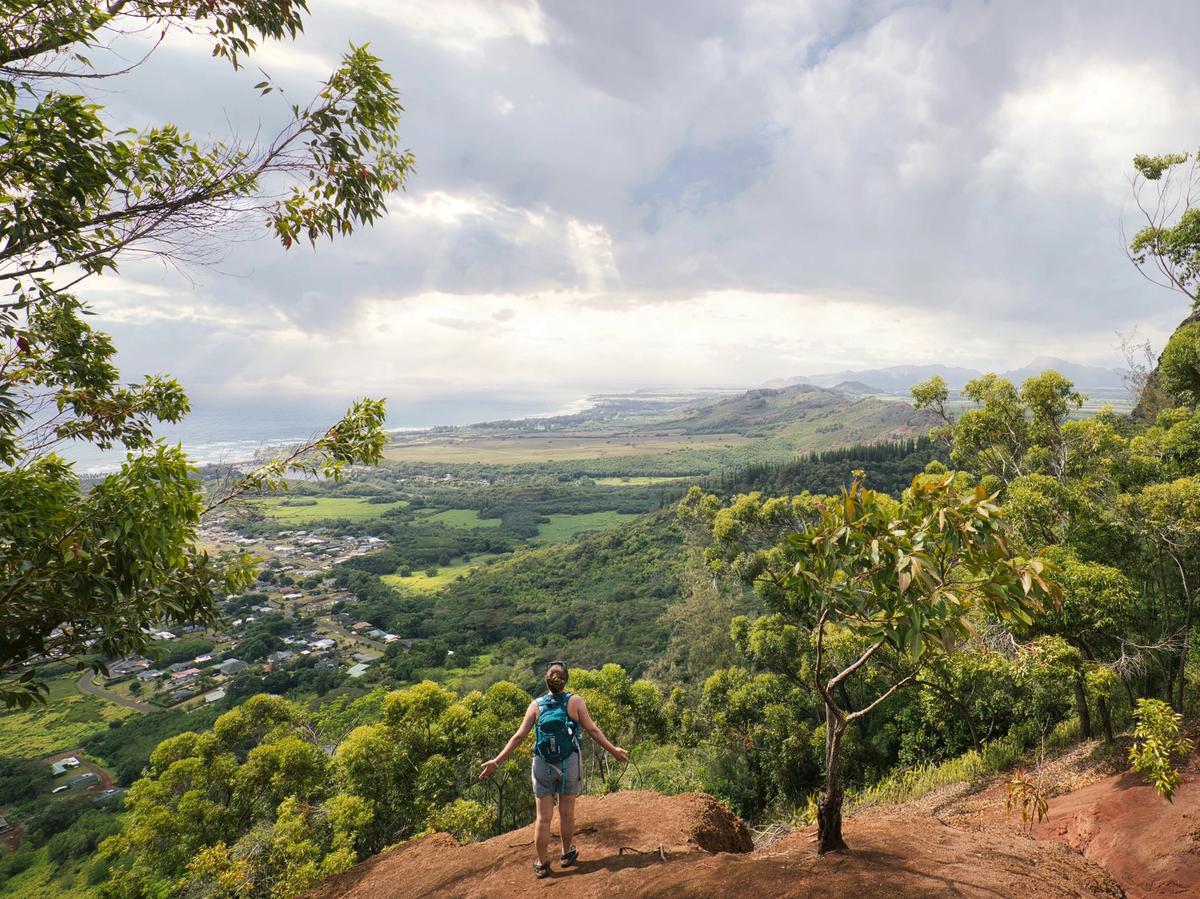 A hiker with arms outstretched stands on a red dirt overlook, gazing across lush green valleys, small towns, and the ocean under a cloudy sky.