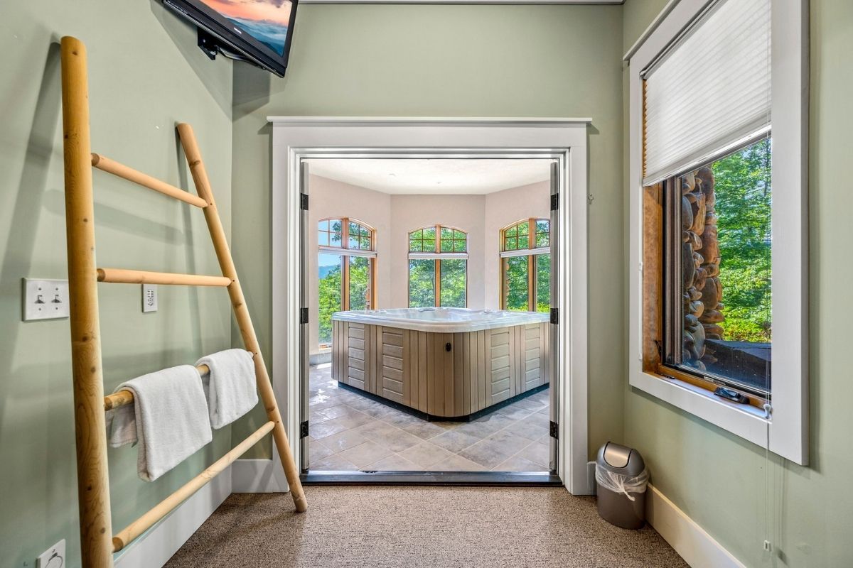 Indoor spa room with a private hot tub framed by large windows overlooking forest views, accessed through double doors, featuring soft green walls, tiled flooring, and a cozy, retreat-style atmosphere.