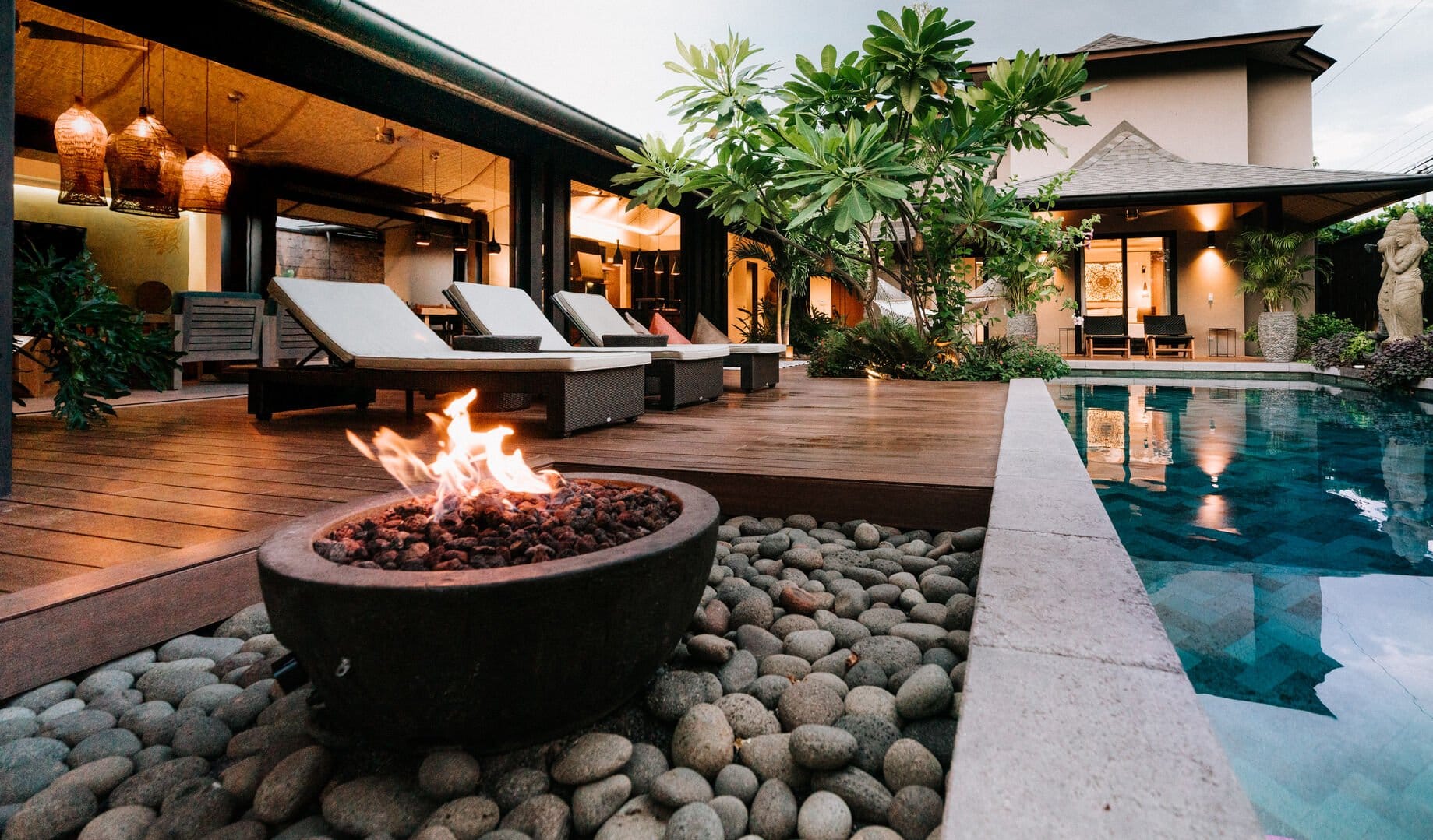 Luxury outdoor space with a stone fire pit, wooden deck loungers, lush greenery, and a serene pool beside a modern villa.