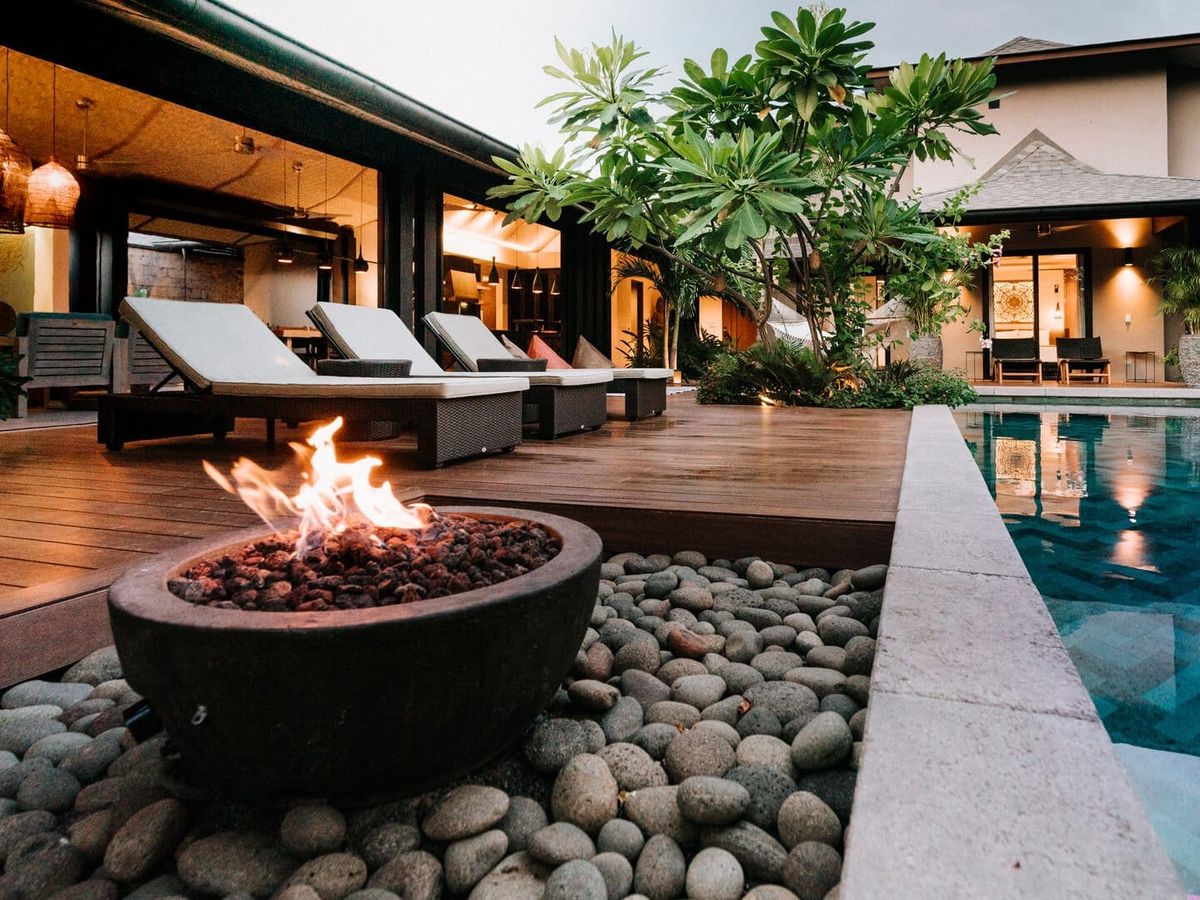 Luxury outdoor space with a stone fire pit, wooden deck loungers, lush greenery, and a serene pool beside a modern villa.