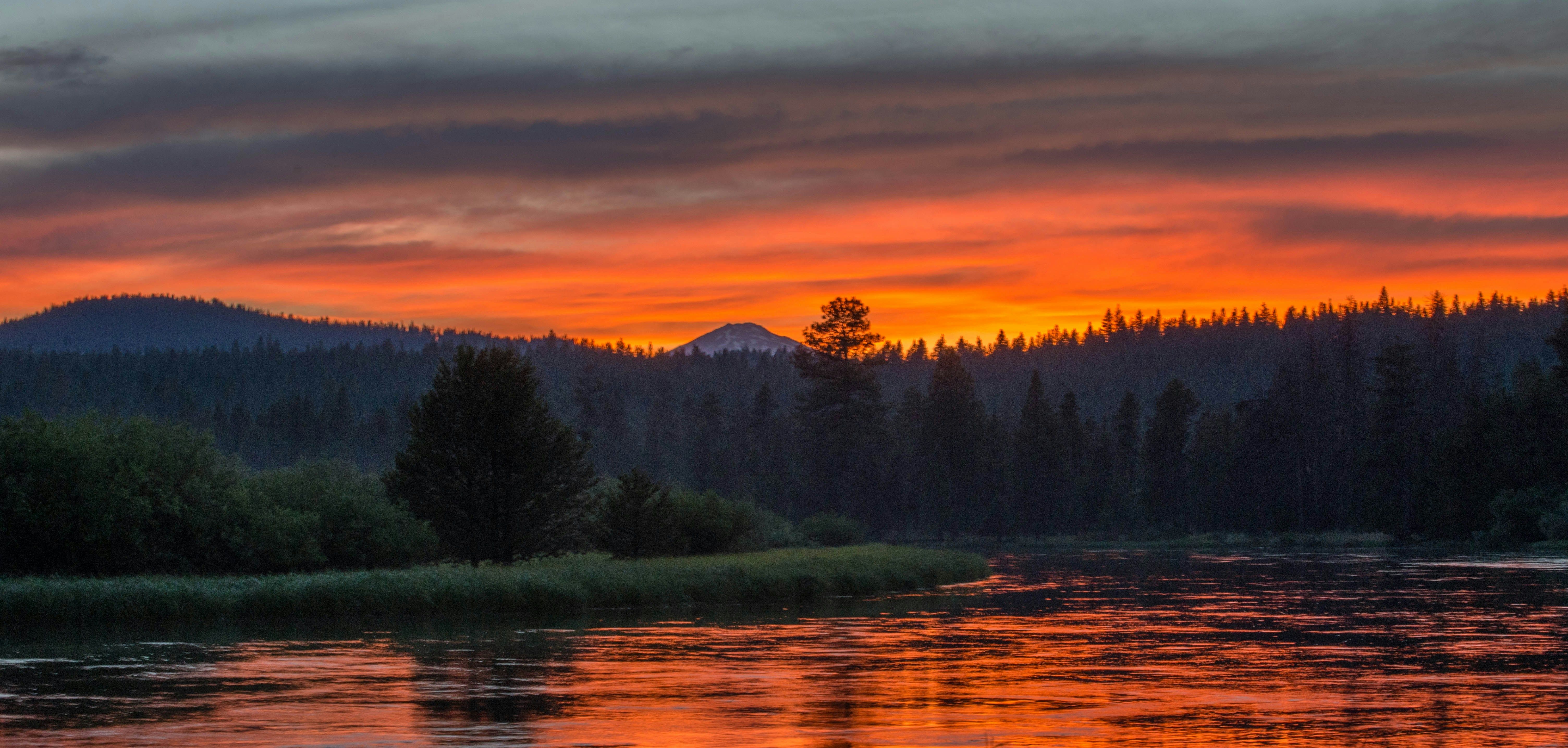 A vibrant sunset over a calm river surrounded by dark pine trees and distant mountains, with the sky glowing in deep shades of orange and red.