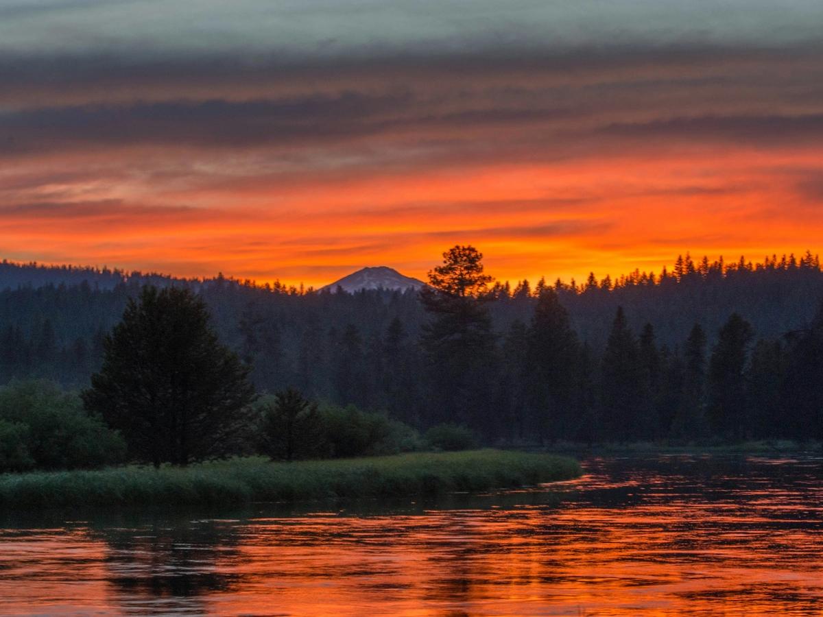 A vibrant sunset over a calm river surrounded by dark pine trees and distant mountains, with the sky glowing in deep shades of orange and red.
