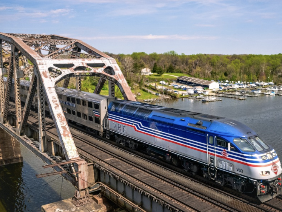 A VRE commuter train crosses a rusted steel bridge over a river, with a marina and lush green trees visible in the background.