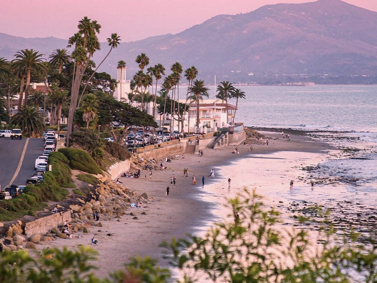 A coastal scene at sunset with palm trees lining the road, people walking along the sandy beach, and mountains in the distance under a pink and purple sky.