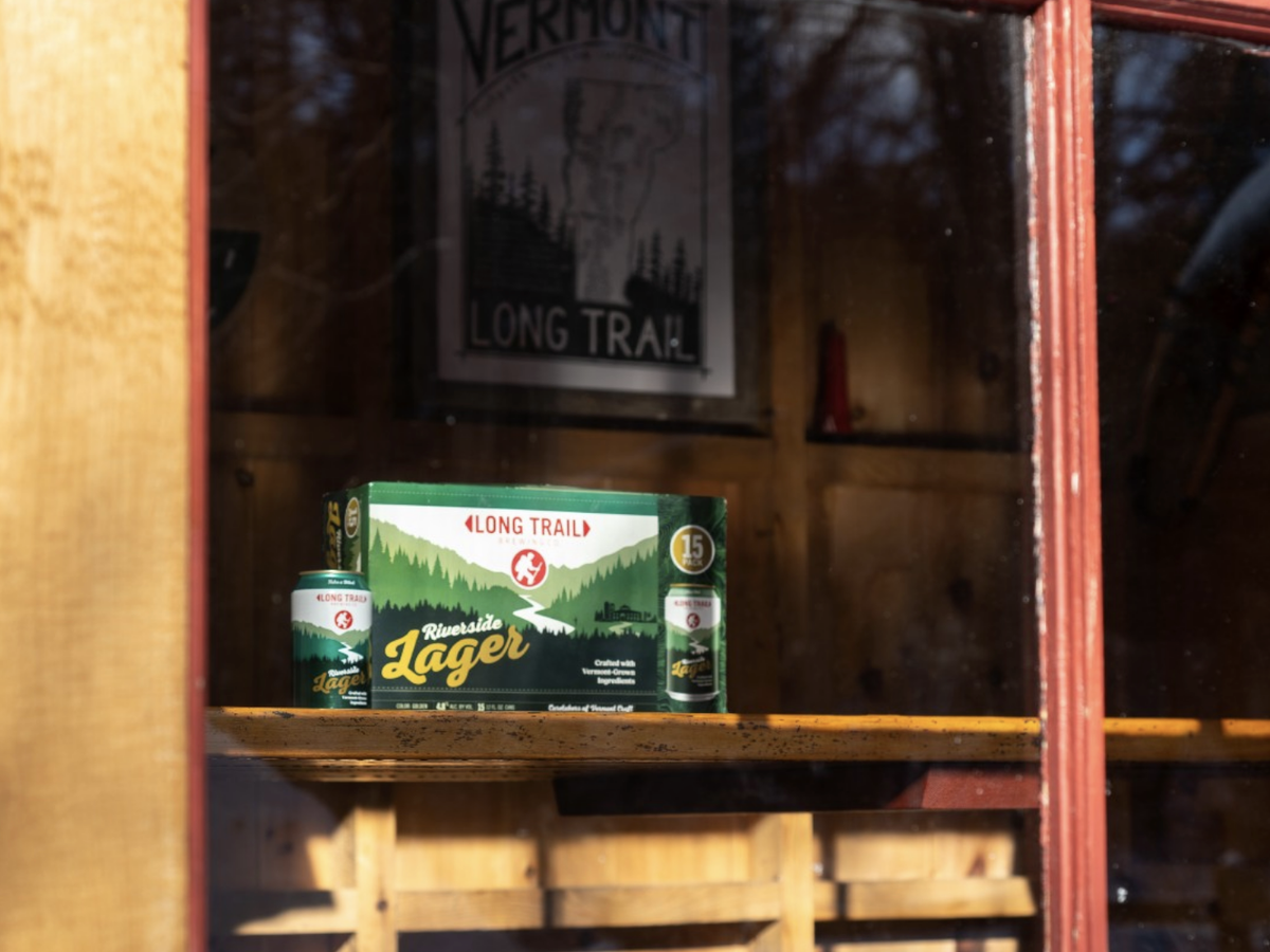 A box and can of Long Trail Riverside Lager sit on a wooden windowsill inside a rustic cabin, with a Vermont Long Trail poster in the background.