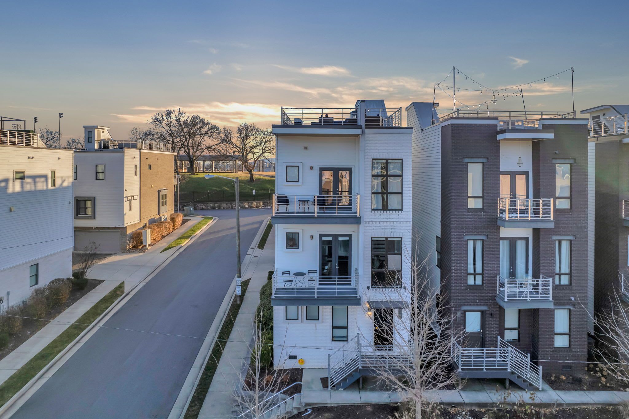 A modern Nashville townhome with multiple balconies and a rooftop deck, set along a quiet residential street at sunset, showcasing sleek architecture and city views ideal for group stays.