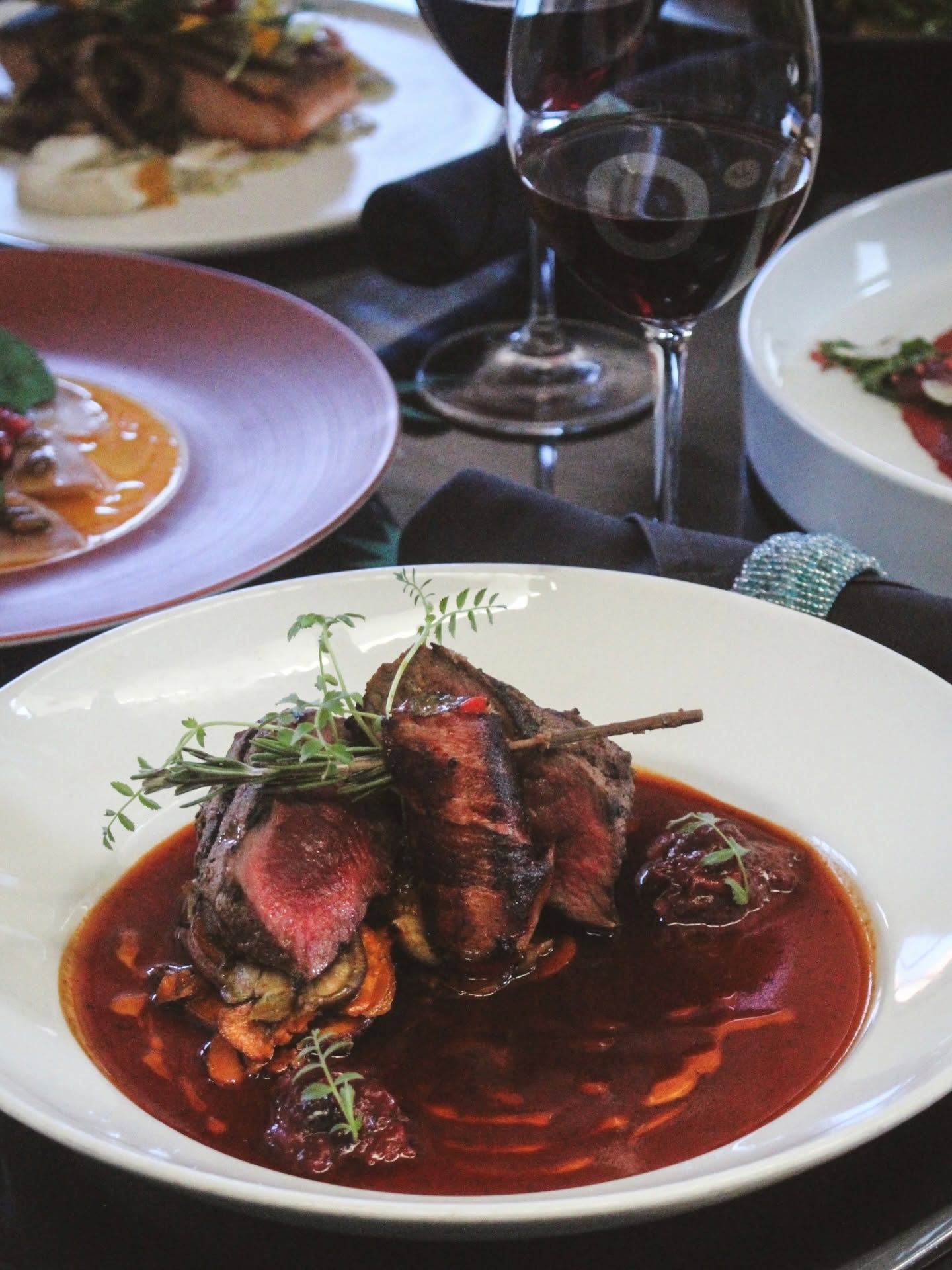 A beautifully plated dish of sliced grilled beef rests over roasted vegetables in a pool of rich red sauce, garnished with fresh herbs on a white plate. A glass of red wine and additional elegantly plated courses fill the softly lit dining table.