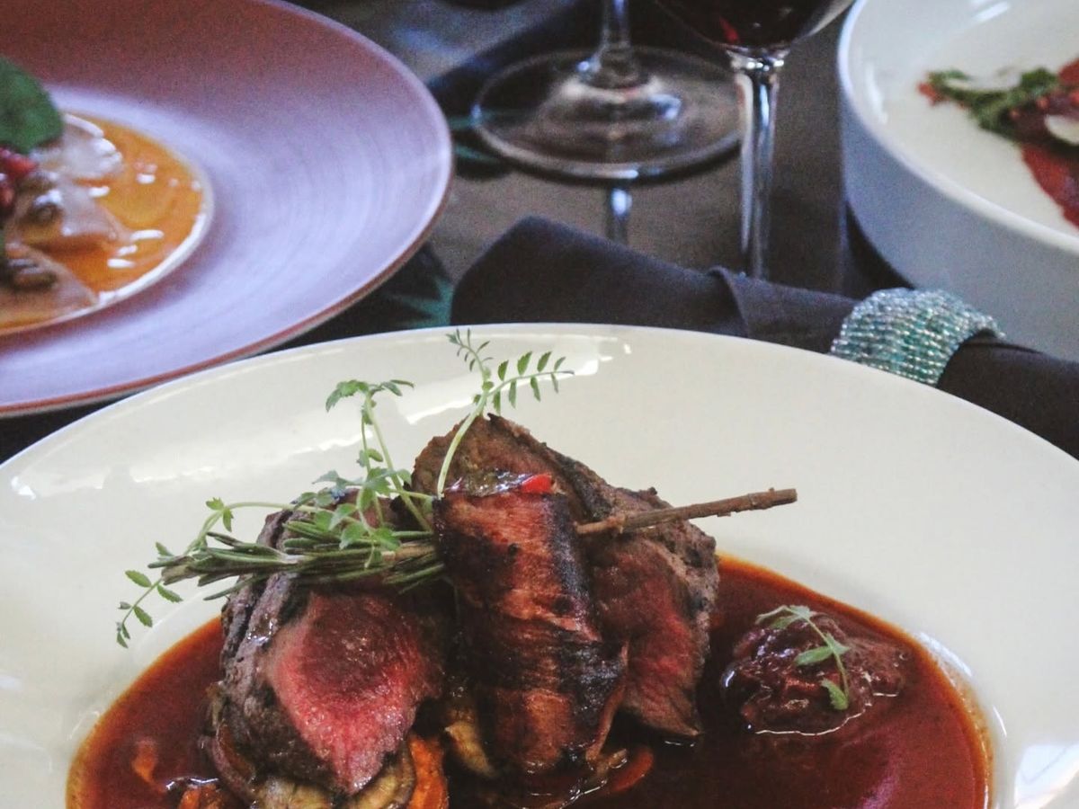 A beautifully plated dish of sliced grilled beef rests over roasted vegetables in a pool of rich red sauce, garnished with fresh herbs on a white plate. A glass of red wine and additional elegantly plated courses fill the softly lit dining table.
