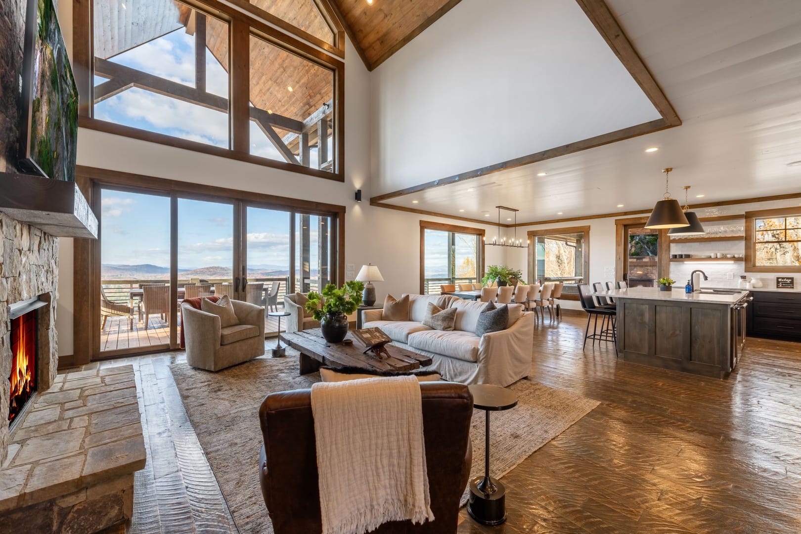 An airy open-concept mountain living space with vaulted wood ceilings, a stone fireplace, expansive windows framing scenic views, a cozy seating area, and a modern kitchen and dining space that open onto a covered deck.