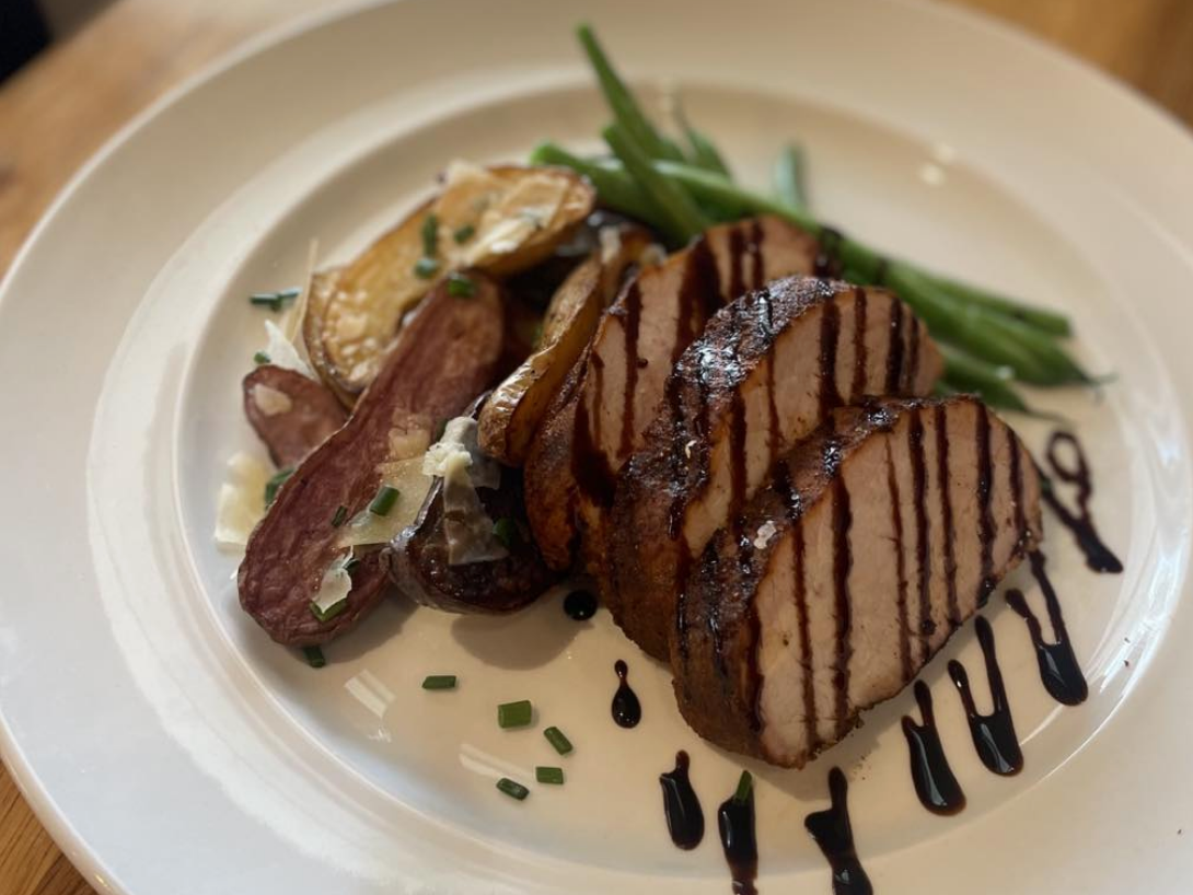 A plated entrée featuring sliced grilled pork tenderloin drizzled with a dark balsamic glaze, served alongside roasted fingerling potatoes with shaved cheese and a portion of fresh green beans.