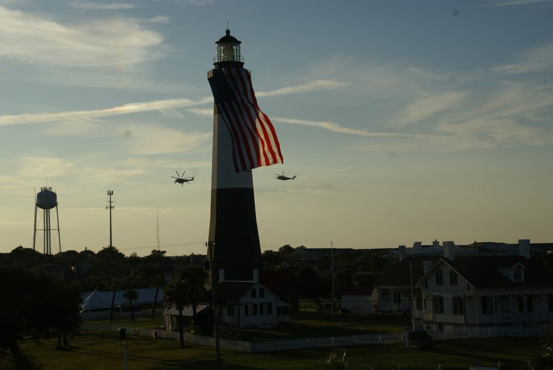 The Tybee Island Lighthouse stands tall with a giant American flag draped across it. Helicopters fly in the background during a dramatic sunset sky, honoring the area’s historic coastal char