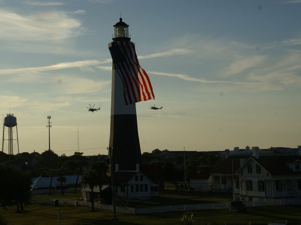 The Tybee Island Lighthouse stands tall with a giant American flag draped across it. Helicopters fly in the background during a dramatic sunset sky, honoring the area’s historic coastal char