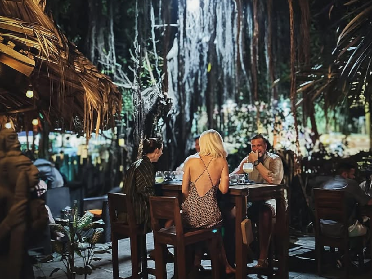 Group of people dining outdoors at a tropical, tiki-style restaurant at night, surrounded by lush trees and hanging vines under soft, ambient lighting