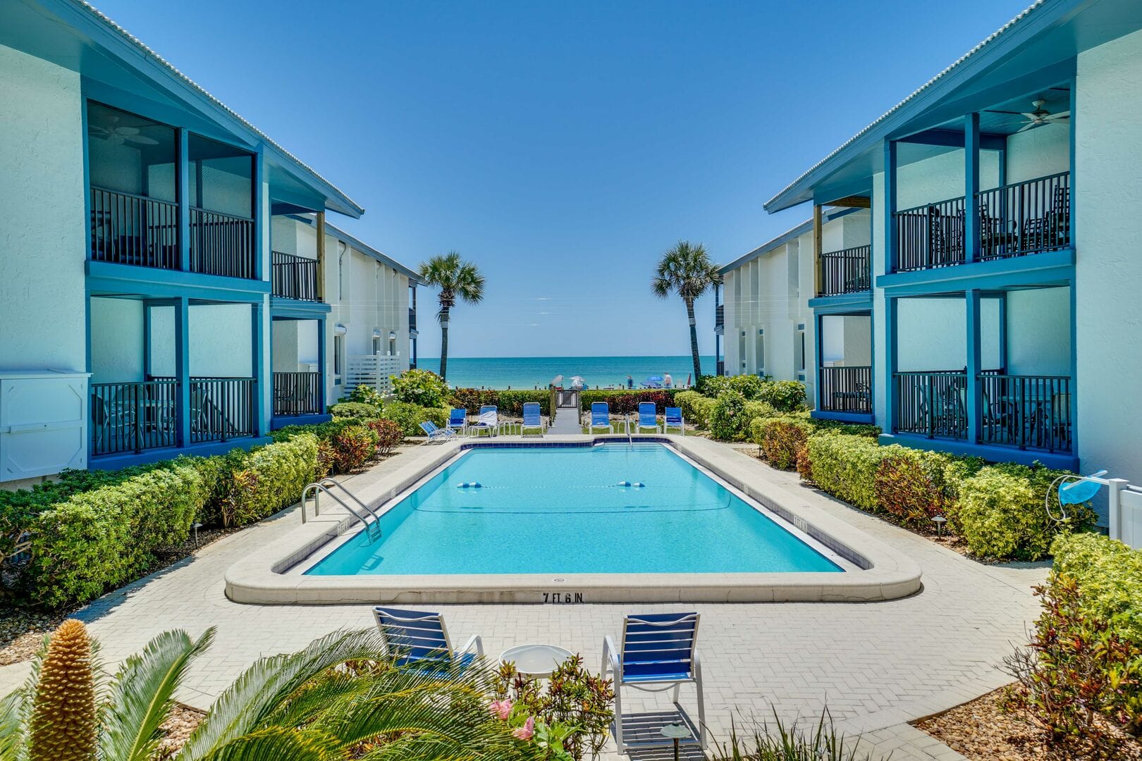 A bright beachfront condo complex features a central swimming pool framed by blue-accented buildings, palm-lined landscaping, and a direct view of the turquoise ocean beyond.