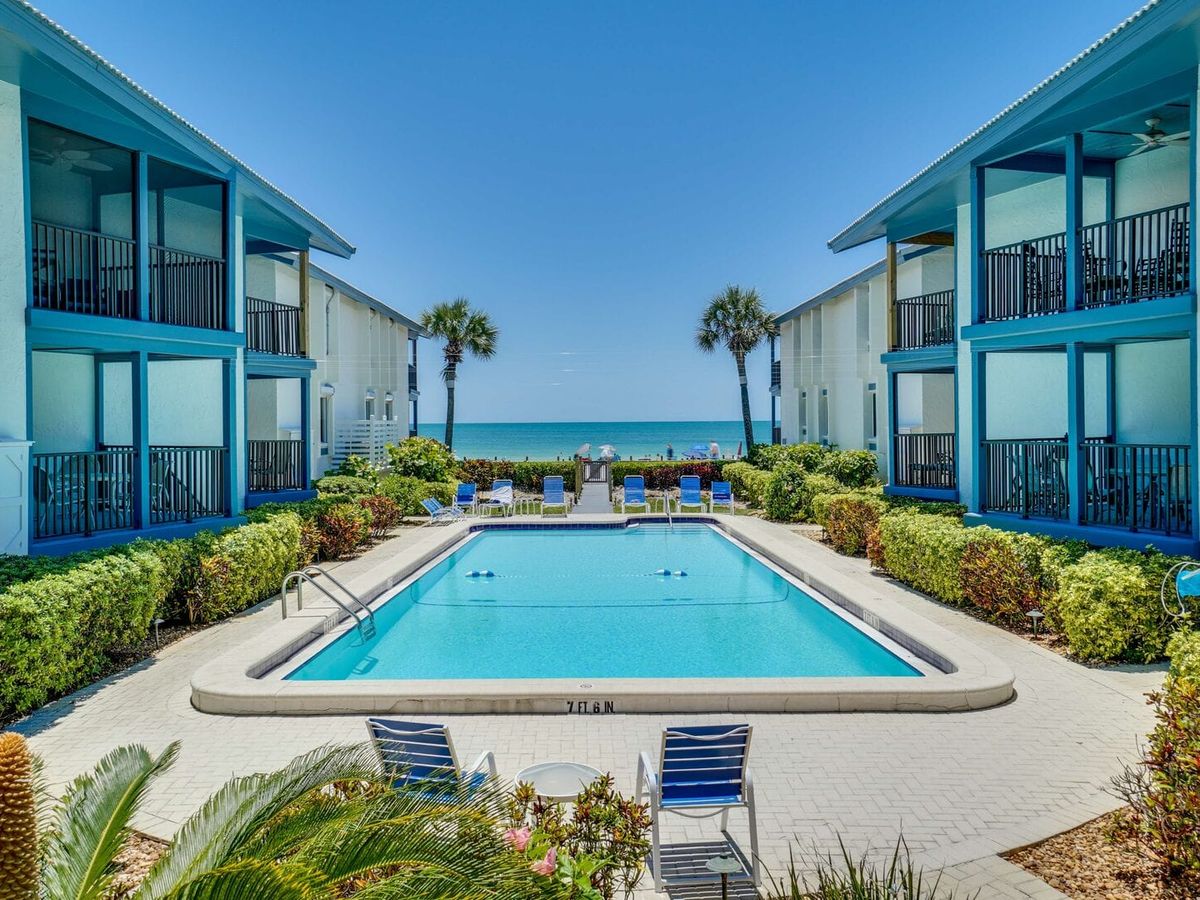 A symmetrical beachfront condo courtyard frames a sparkling rectangular pool with lounge chairs, flanked by turquoise-accented buildings and palm trees, opening to a clear blue ocean horizon under a cloudless sky.