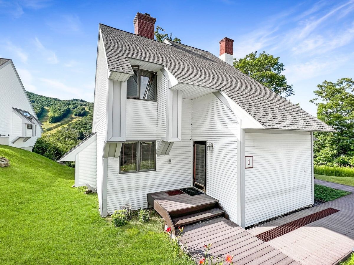 A modern white mountain chalet with steep rooflines sits on a green slope, overlooking scenic ski trails and forested hills in the background.