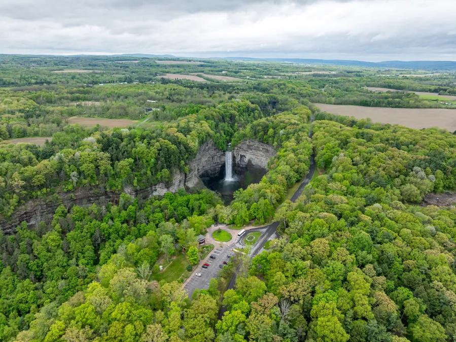 An aerial view of Taughannock Falls State Park showing a tall waterfall dropping into a forested gorge, with winding roads and lush greenery below. This view highlights the scale of one of the tallest waterfalls east of the Rockies.
