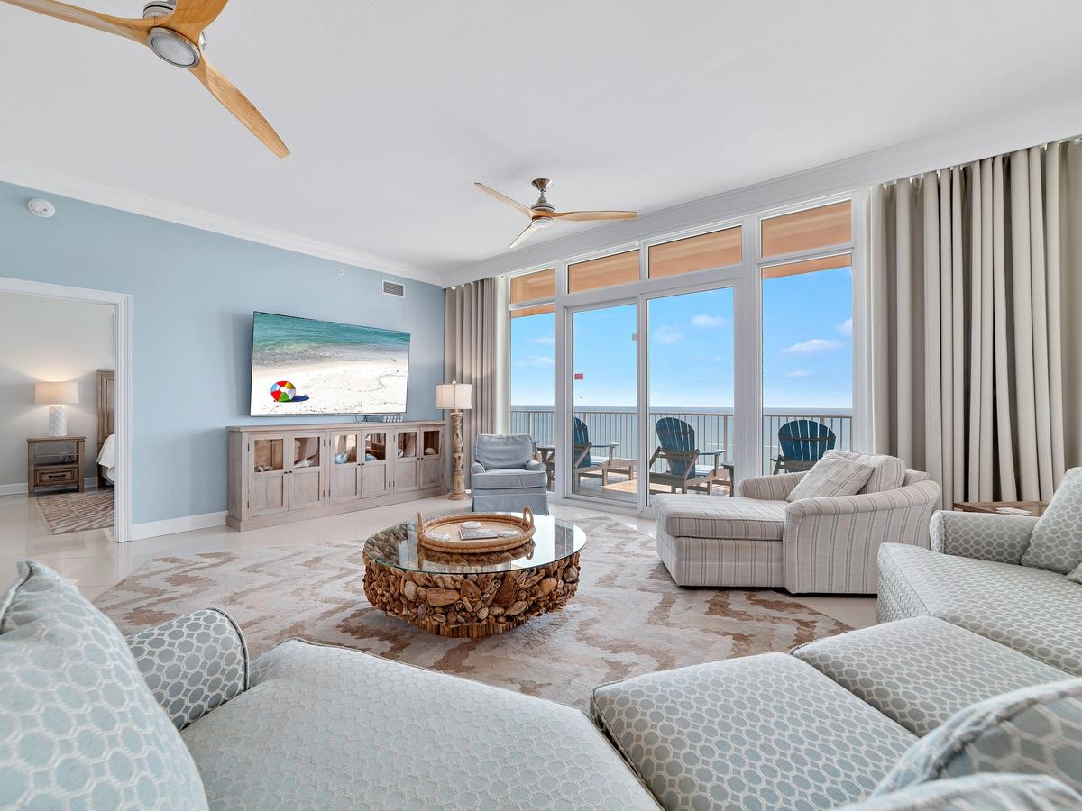 A bright and airy coastal living room with pastel blue walls, light patterned furniture, a driftwood coffee table, and floor-to-ceiling windows that open to a balcony with ocean views and Adirondack chairs.