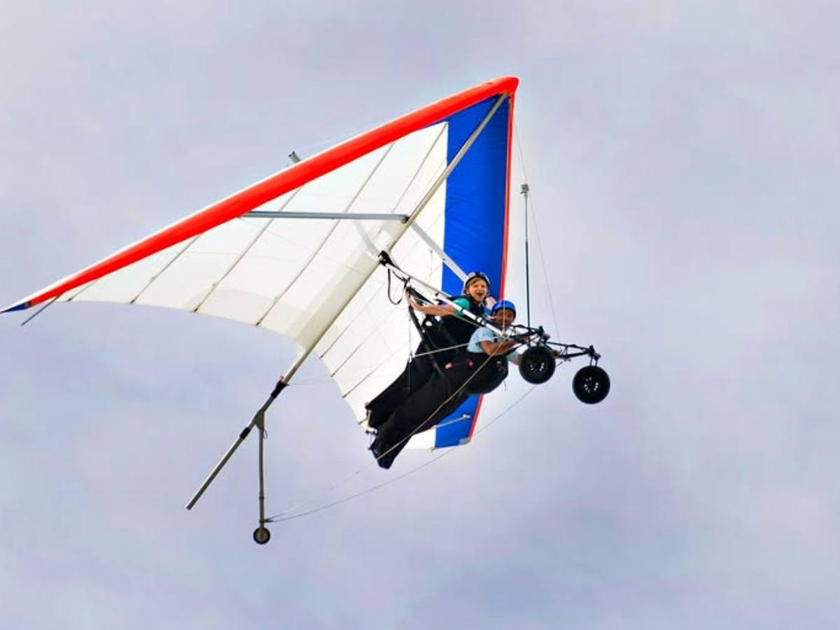 Two people tandem hang gliding under a red, white, and blue wing, soaring high against a cloudy sky.