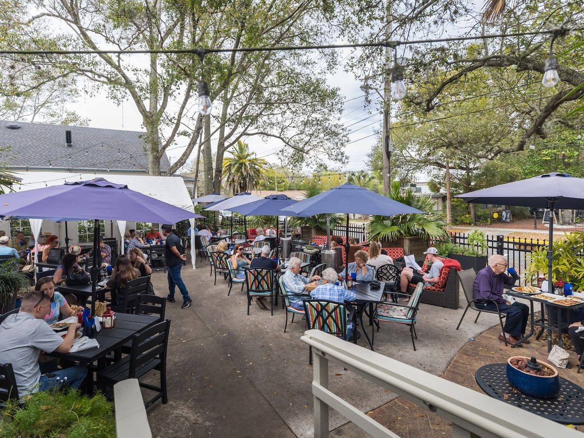 A lively outdoor dining patio filled with guests enjoying meals under large blue umbrellas. String lights hang above, adding charm to the lush, tree-lined setting. The space features a mix of tables and cushioned seating, creating a welcoming and vibrant atmosphere