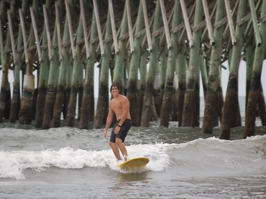 A smiling surfer rides a small wave on a yellow surfboard near a weathered pier with green, algae-covered wooden beams.