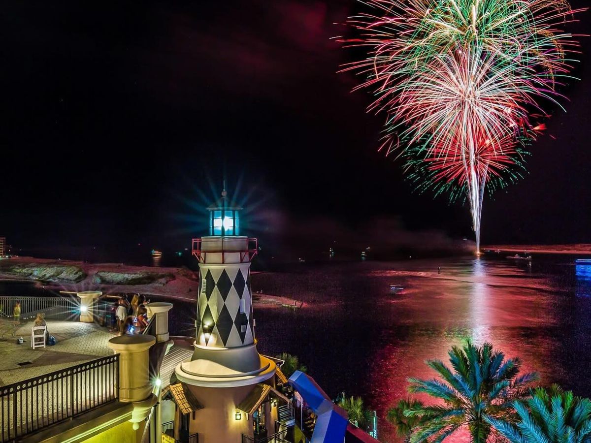 Colorful fireworks light up the night sky over the Destin Harbor, reflecting across the water near the HarborWalk lighthouse. People gather along the waterfront to watch the celebration, creating a lively New Year’s Eve scene by the marina. This image captures the excitement of fireworks over Destin’s harbor at night.