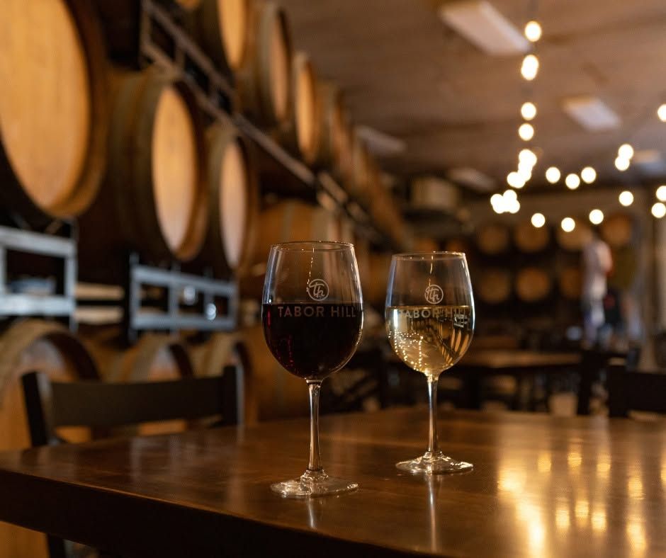 Two Tabor Hill wine glasses, one filled with red and one with white, rest on a polished wooden table inside a warmly lit barrel room. Rows of oak wine barrels and glowing string lights create an intimate, rustic tasting atmosphere. 