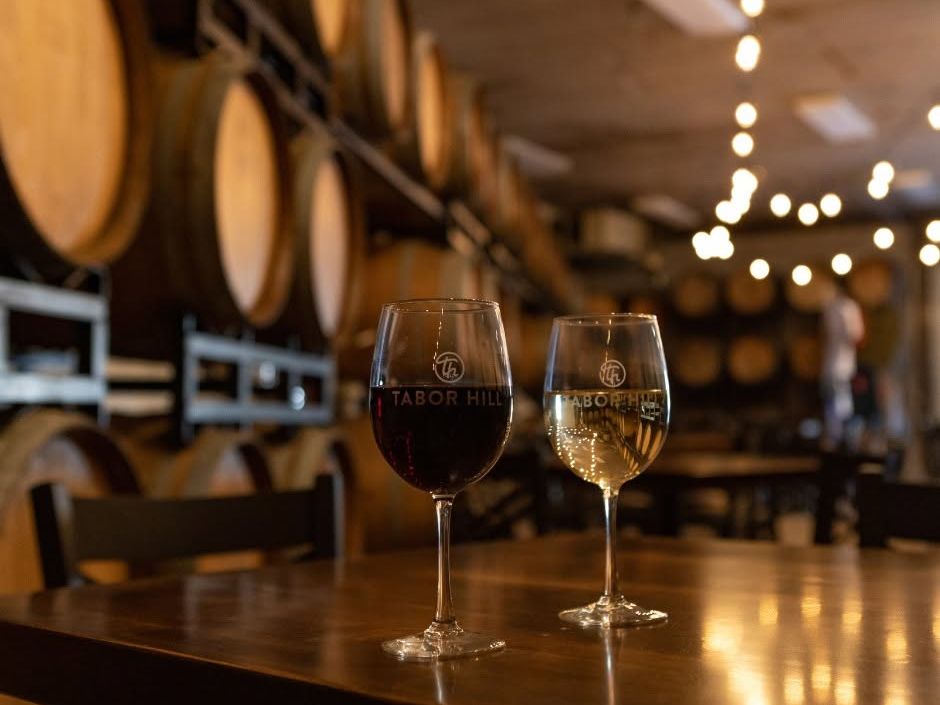 Two Tabor Hill wine glasses, one filled with red and one with white, rest on a polished wooden table inside a warmly lit barrel room. Rows of oak wine barrels and glowing string lights create an intimate, rustic tasting atmosphere.