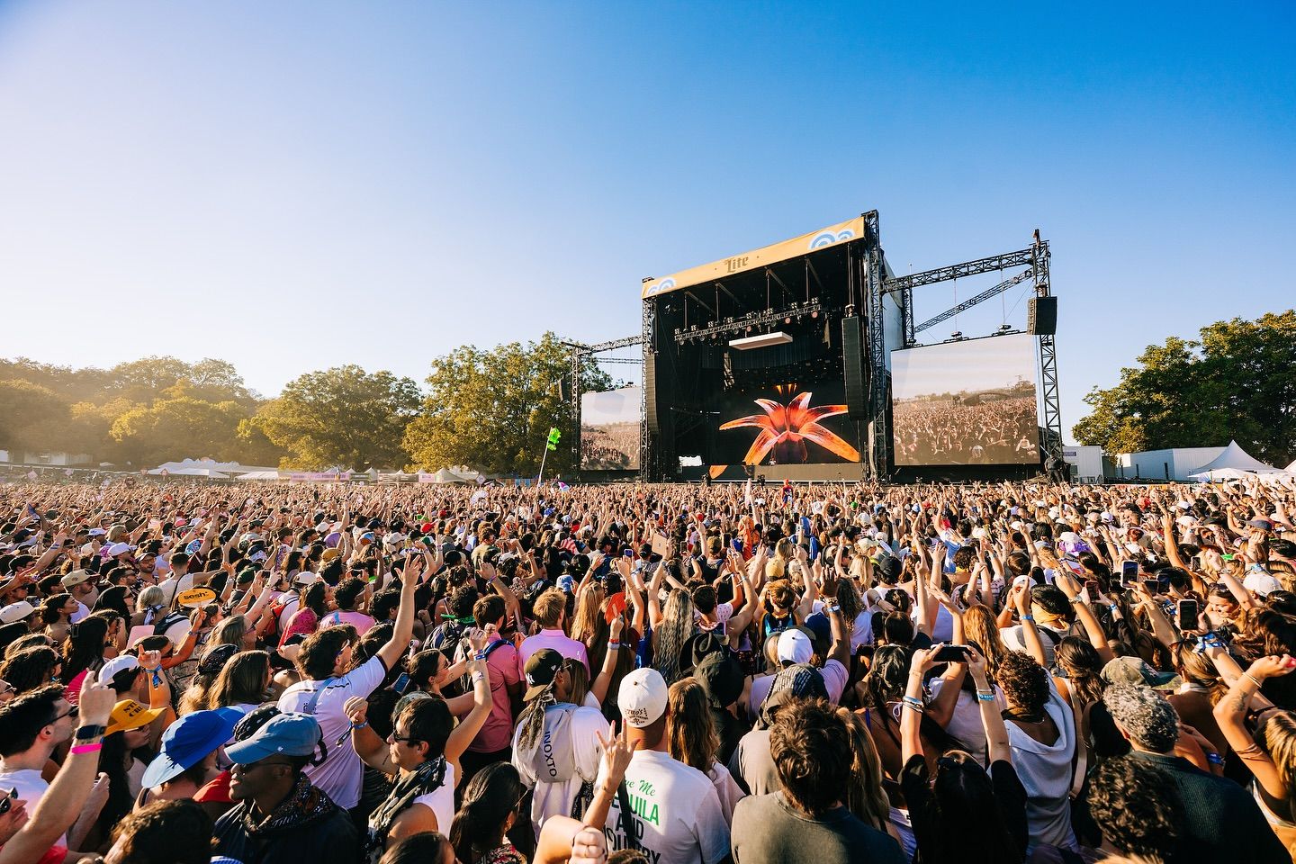 A massive festival crowd raises their hands toward the main stage, soaking in the music under a clear blue Texas sky. The energy feels electric as thousands of fans enjoy the outdoor concert together. Bright screens and stage lights make the moment feel larger than life.