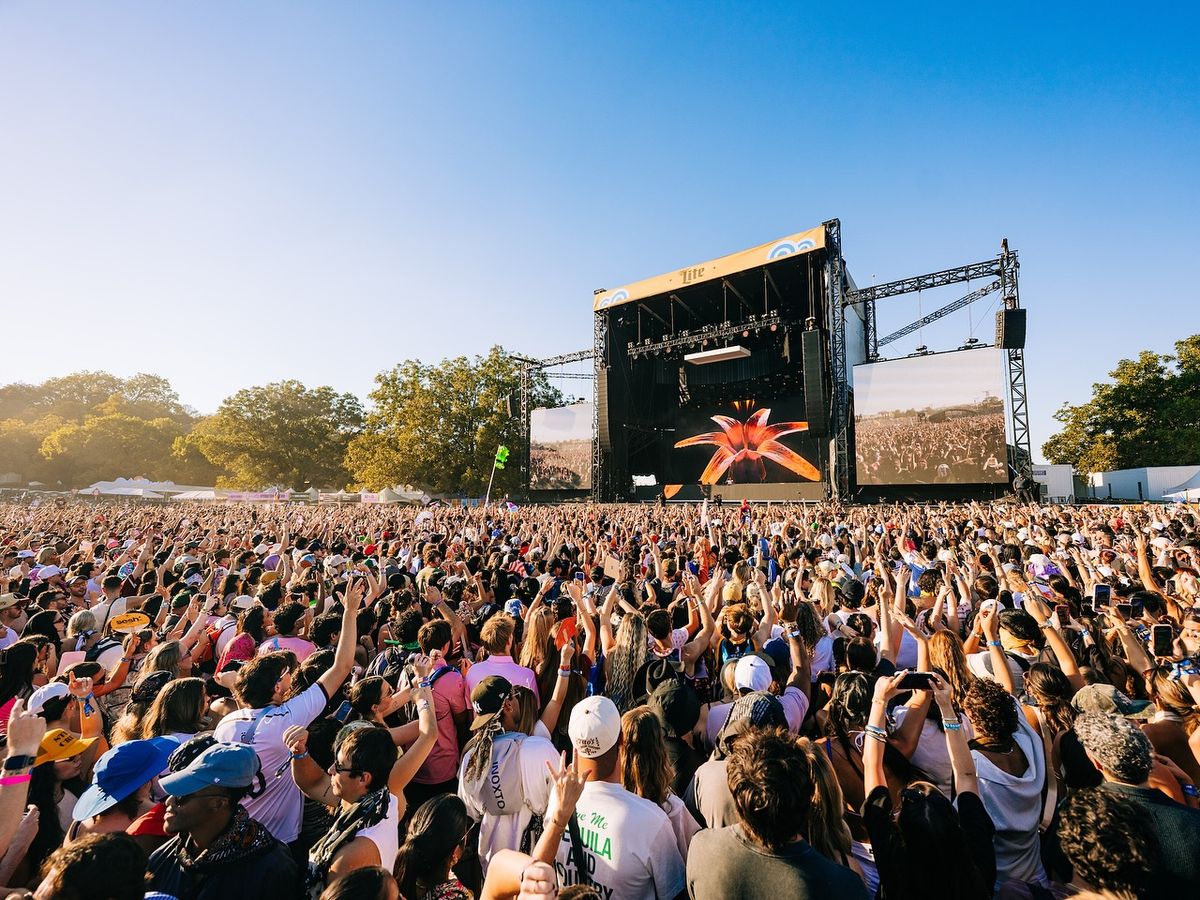 A massive festival crowd raises their hands toward the main stage, soaking in the music under a clear blue Texas sky. The energy feels electric as thousands of fans enjoy the outdoor concert together. Bright screens and stage lights make the moment feel larger than life.