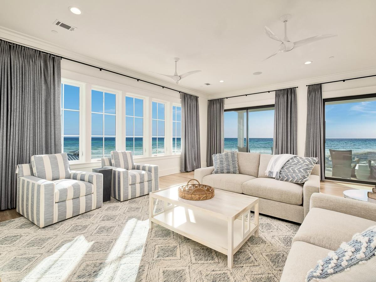 A bright coastal living room with ocean views, featuring neutral-toned sofas, striped armchairs, and large windows that frame the turquoise water and blue sky.