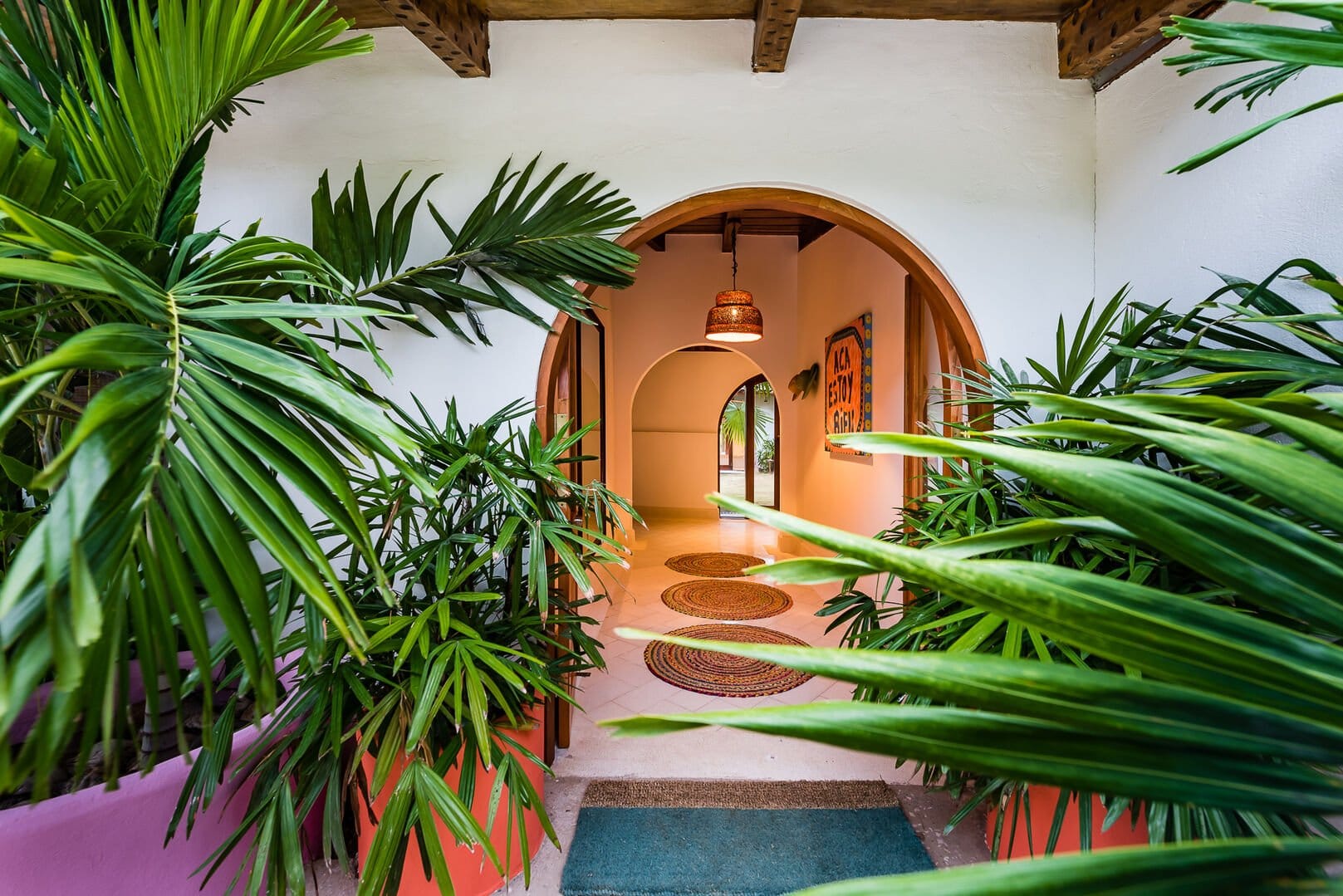 Colorful tropical entryway with lush potted plants, arched doorways, woven rugs, and warm accent lighting.