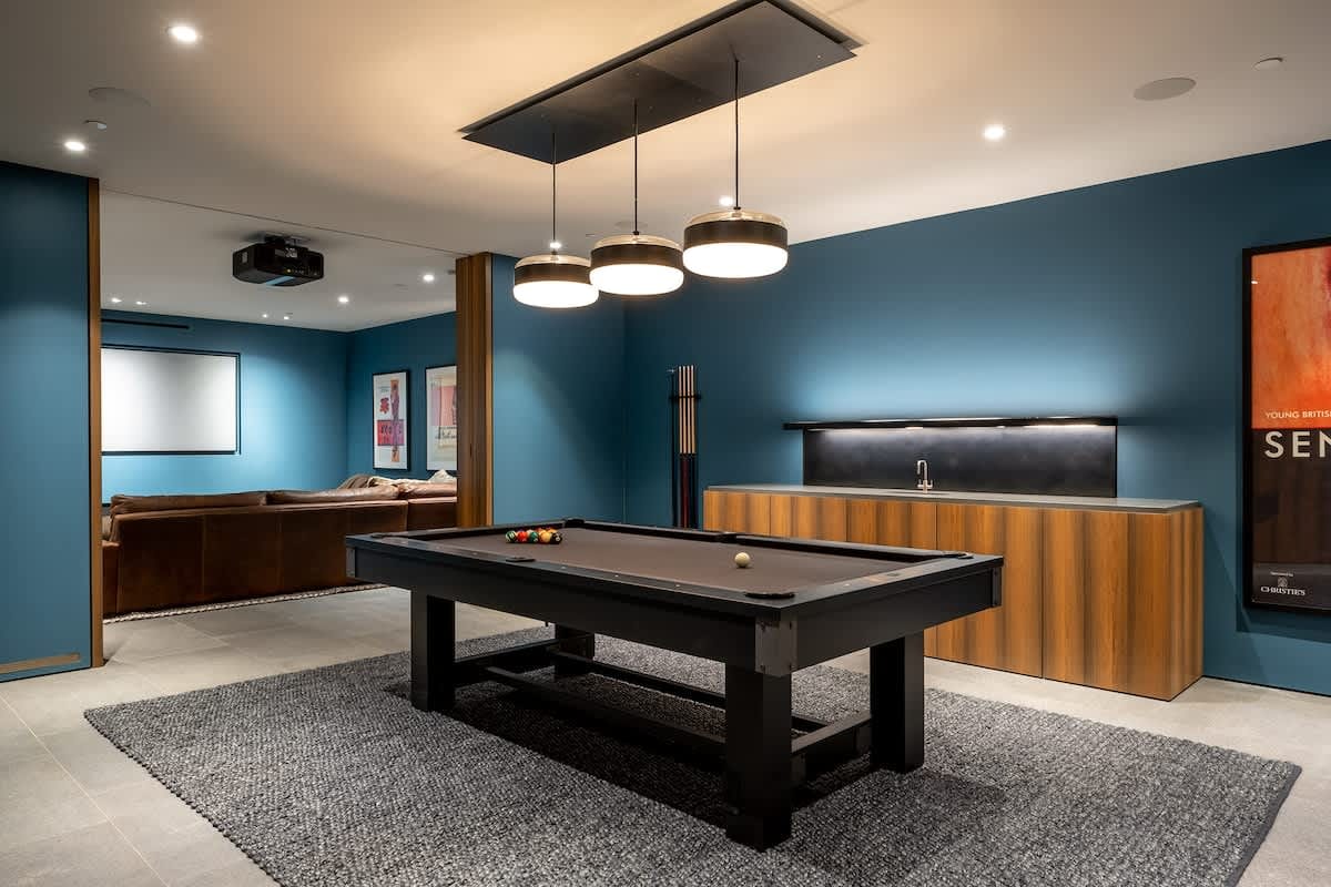 Contemporary game room with a sleek pool table on a textured rug, teal accent walls, modern pendant lighting, a built-in wet bar, and a cozy lounge area with a projector screen in the background.