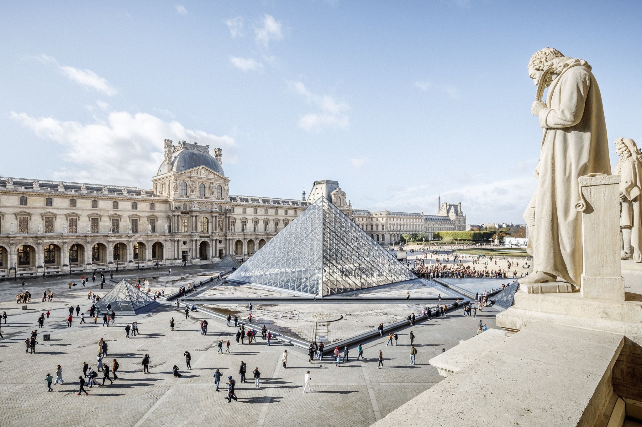 A wide view of the Louvre shows the famous glass pyramid surrounded by people walking across the grand courtyard. The historic stone buildings frame the modern structure beautifully under a bright blue sky. It captures the perfect mix of old-world Paris and modern design.