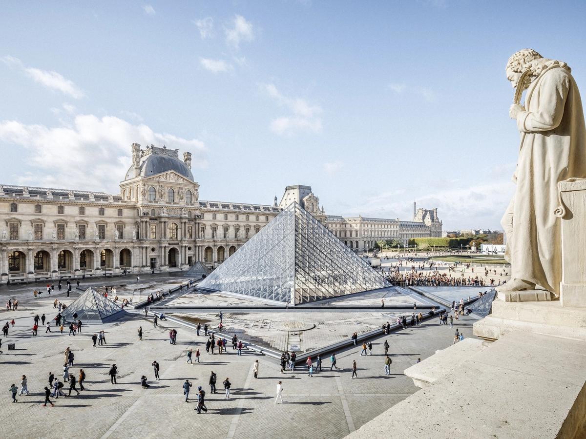 A wide view of the Louvre shows the famous glass pyramid surrounded by people walking across the grand courtyard. The historic stone buildings frame the modern structure beautifully under a bright blue sky. It captures the perfect mix of old-world Paris and modern design.