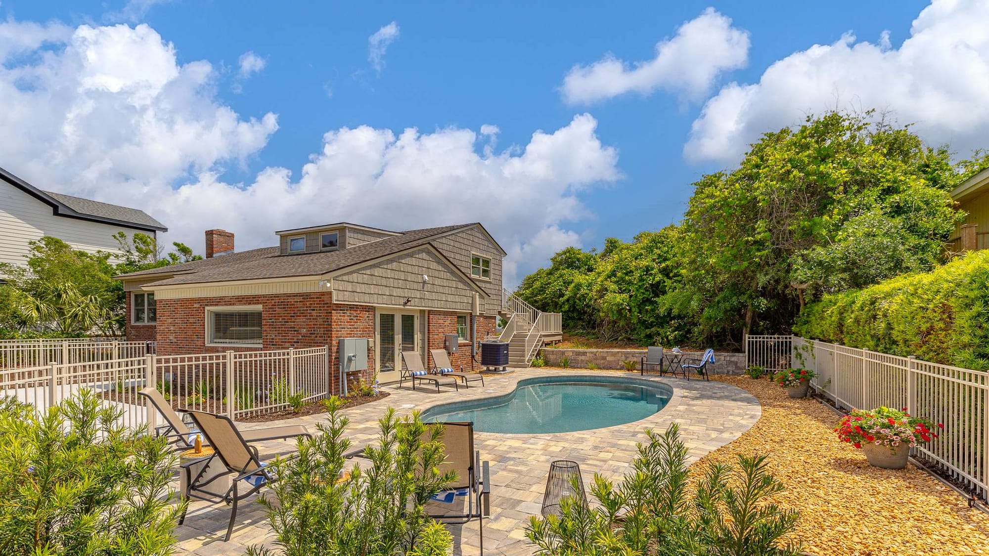 The backyard of this brick beach home features a freeform pool surrounded by a paver patio with lounge chairs, framed by lush landscaping and a white picket fence. A second-story deck with staircase access and a dormer window overlook the private outdoor space, perfect for a family vacation rental.