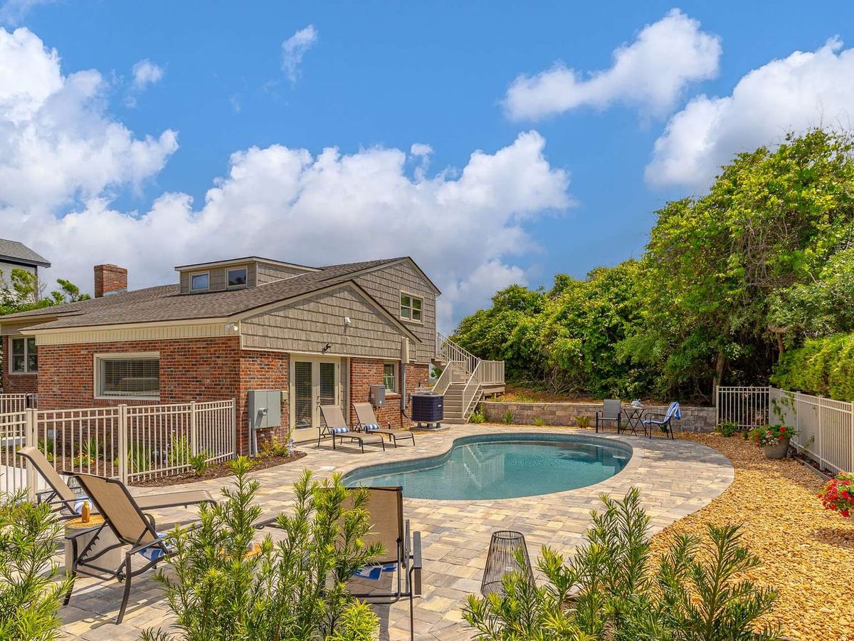 The backyard of this brick beach home features a freeform pool surrounded by a paver patio with lounge chairs, framed by lush landscaping and a white picket fence. A second-story deck with staircase access and a dormer window overlook the private outdoor space, perfect for a family vacation rental.