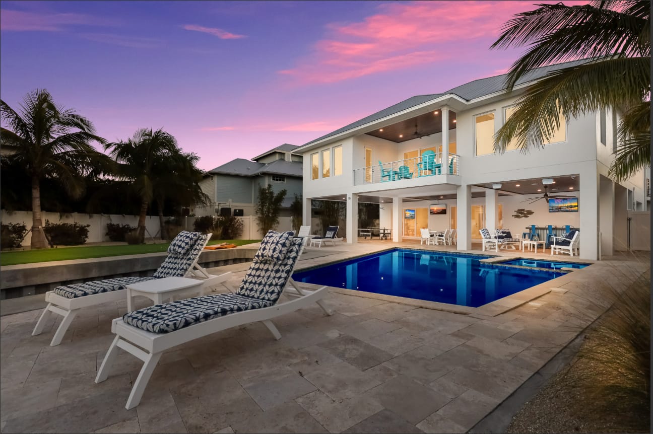 A modern Florida vacation home on Anna Maria Island with a sparkling blue pool and two lounge chairs set out on a travertine deck under a vivid pink and purple sunset sky. Palm trees frame the two-story white house, which has a covered upper balcony with teal chairs and a fully lit outdoor living area below