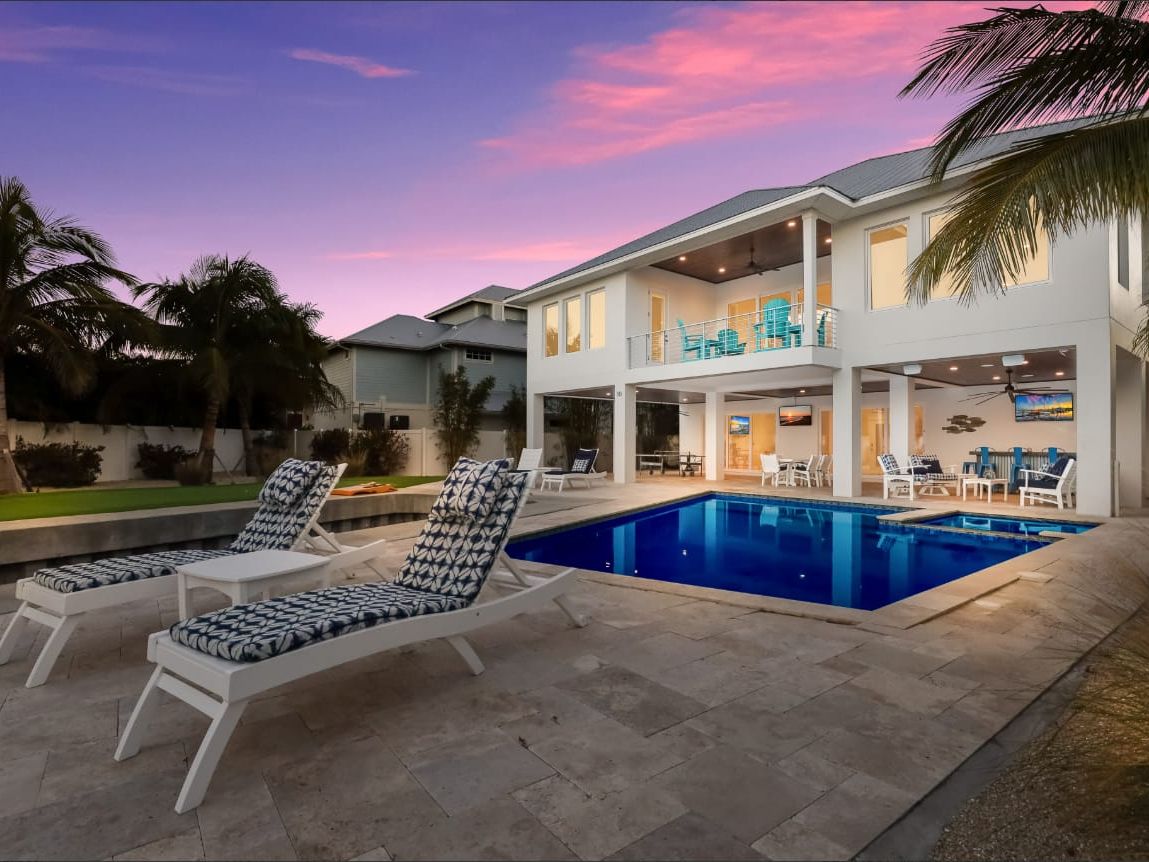 A modern Florida vacation home on Anna Maria Island with a sparkling blue pool and two lounge chairs set out on a travertine deck under a vivid pink and purple sunset sky. Palm trees frame the two-story white house, which has a covered upper balcony with teal chairs and a fully lit outdoor living area below