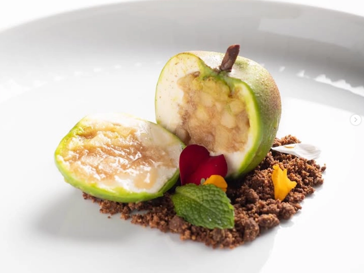 Gourmet dessert styled to look like a green pear, served with edible soil, flower petals, and mint on a white plate.