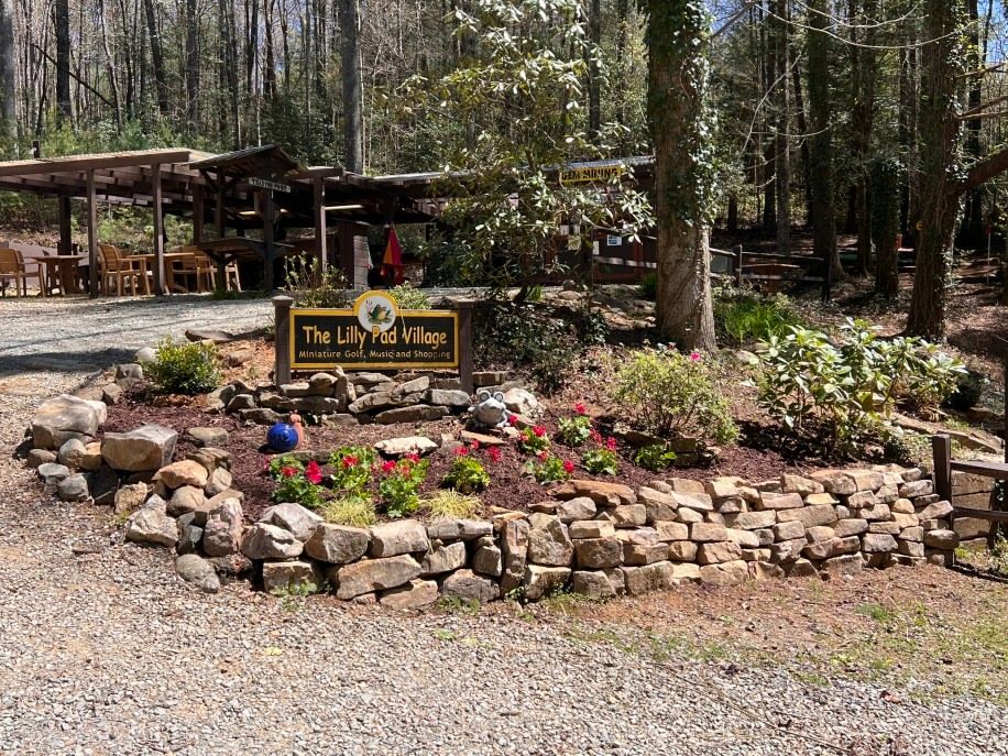 The Lilly Pad Village sign welcomes guests to a charming outdoor attraction surrounded by tall trees. Bright flowers, stone landscaping, and rustic wooden seating create a cozy, family-friendly vibe. It’s the perfect spot for mini golf, gem mining, and mountain fun.