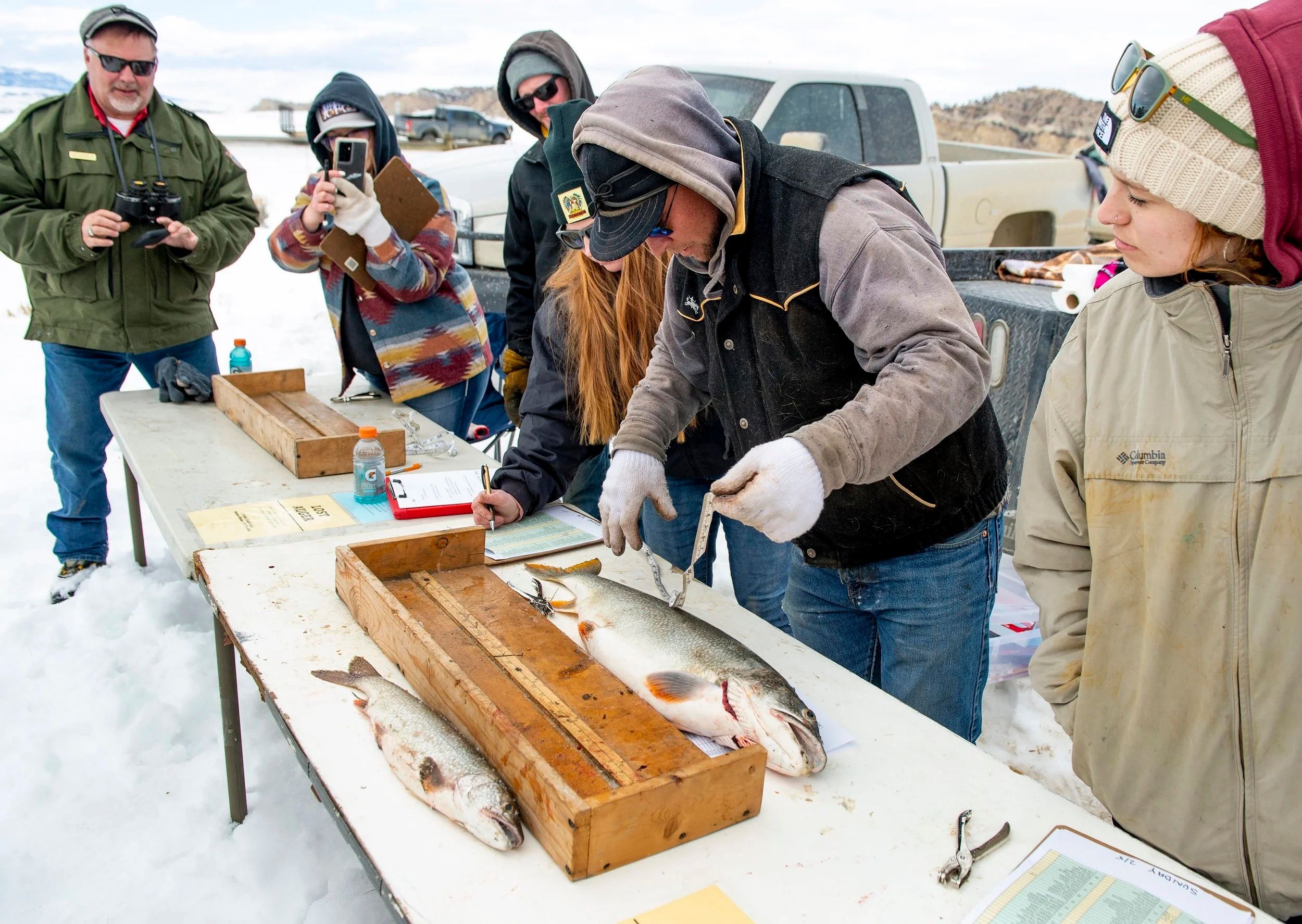 Participants gather around tables in the snow, measuring and recording their catches during the Meeteetse Ice Fishing Derby. The friendly competition brings together locals and anglers to enjoy a crisp winter day outdoors.
