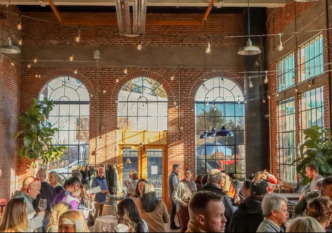 A lively indoor gathering in a brick-walled venue with tall arched windows, string lights, and guests mingling with drinks.